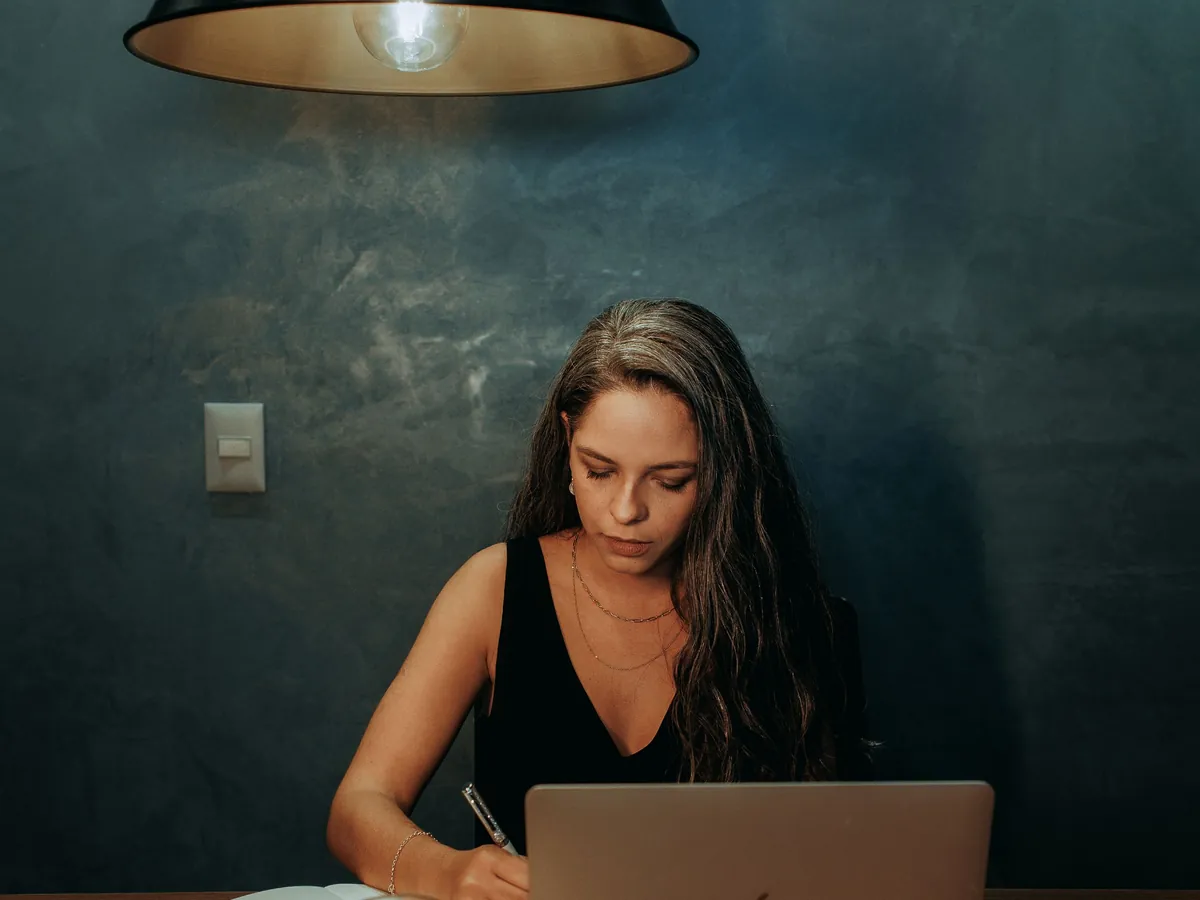 An adult writing notes beside a laptop. Illustrative stock photography, not a customer portrait.